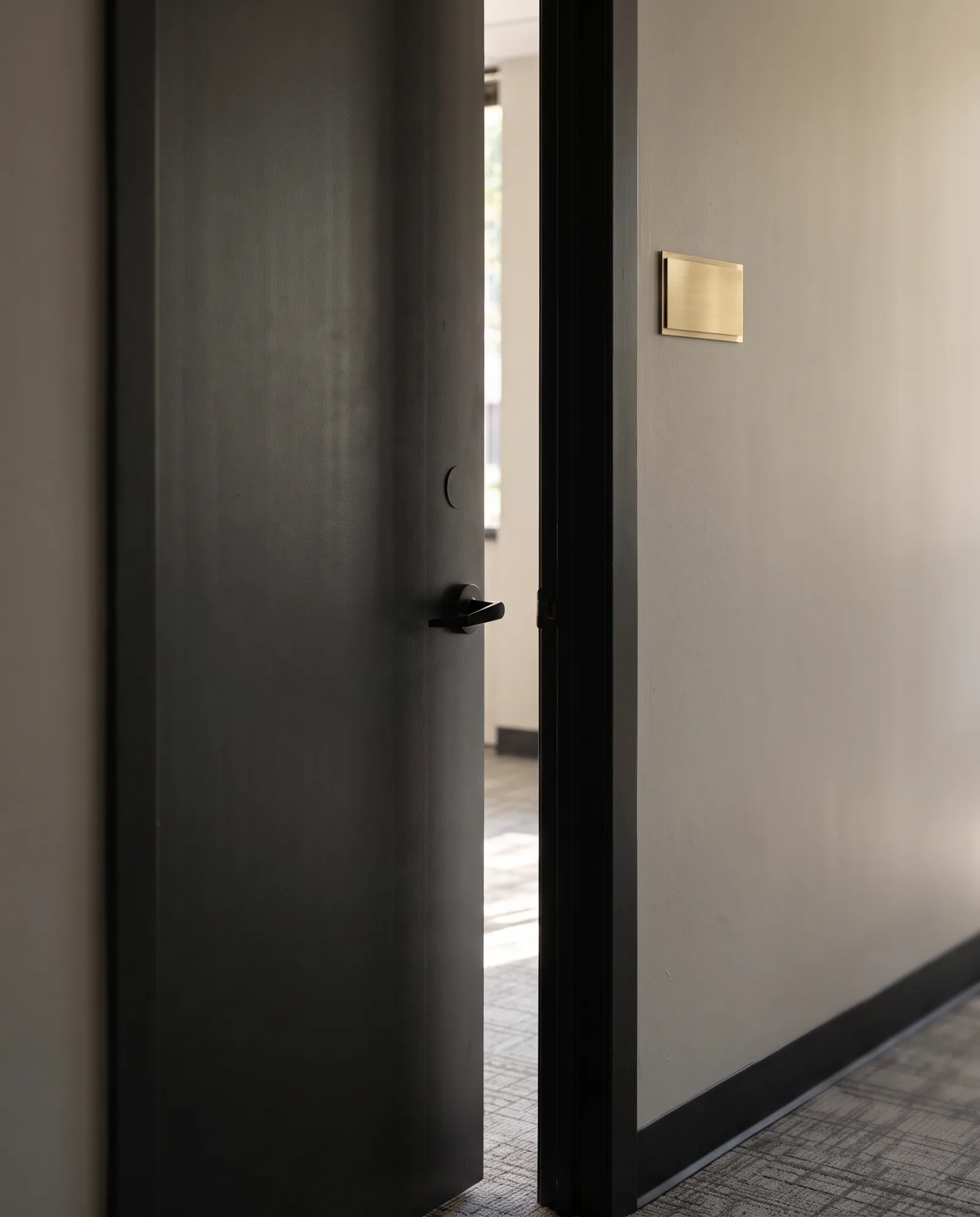 Staged photo: a dark office door with a blank brass nameplate beside it, slightly ajar onto a sunlit room