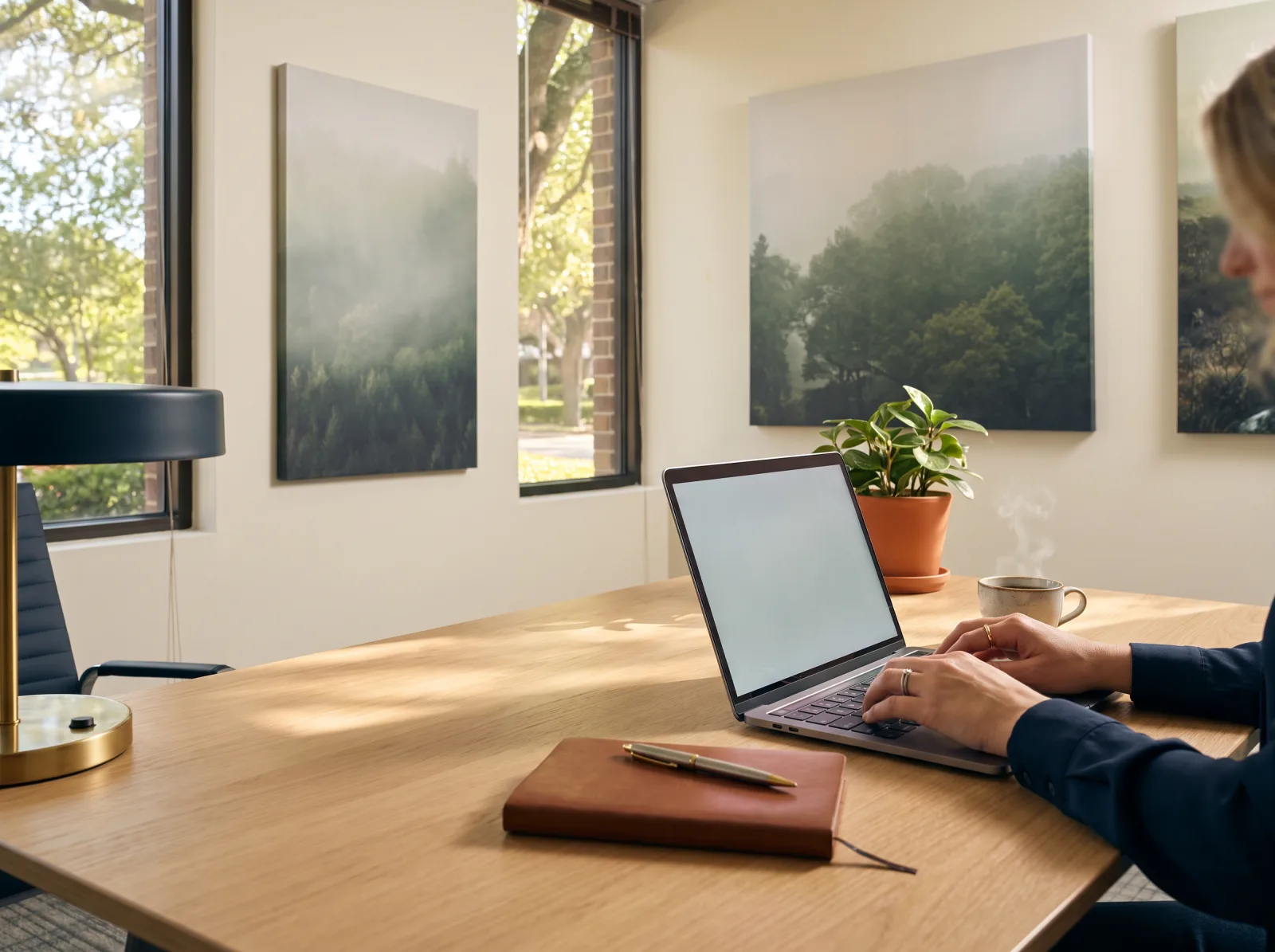 Staged photo: a laptop, notebook and coffee on an oak table by a window onto live oaks