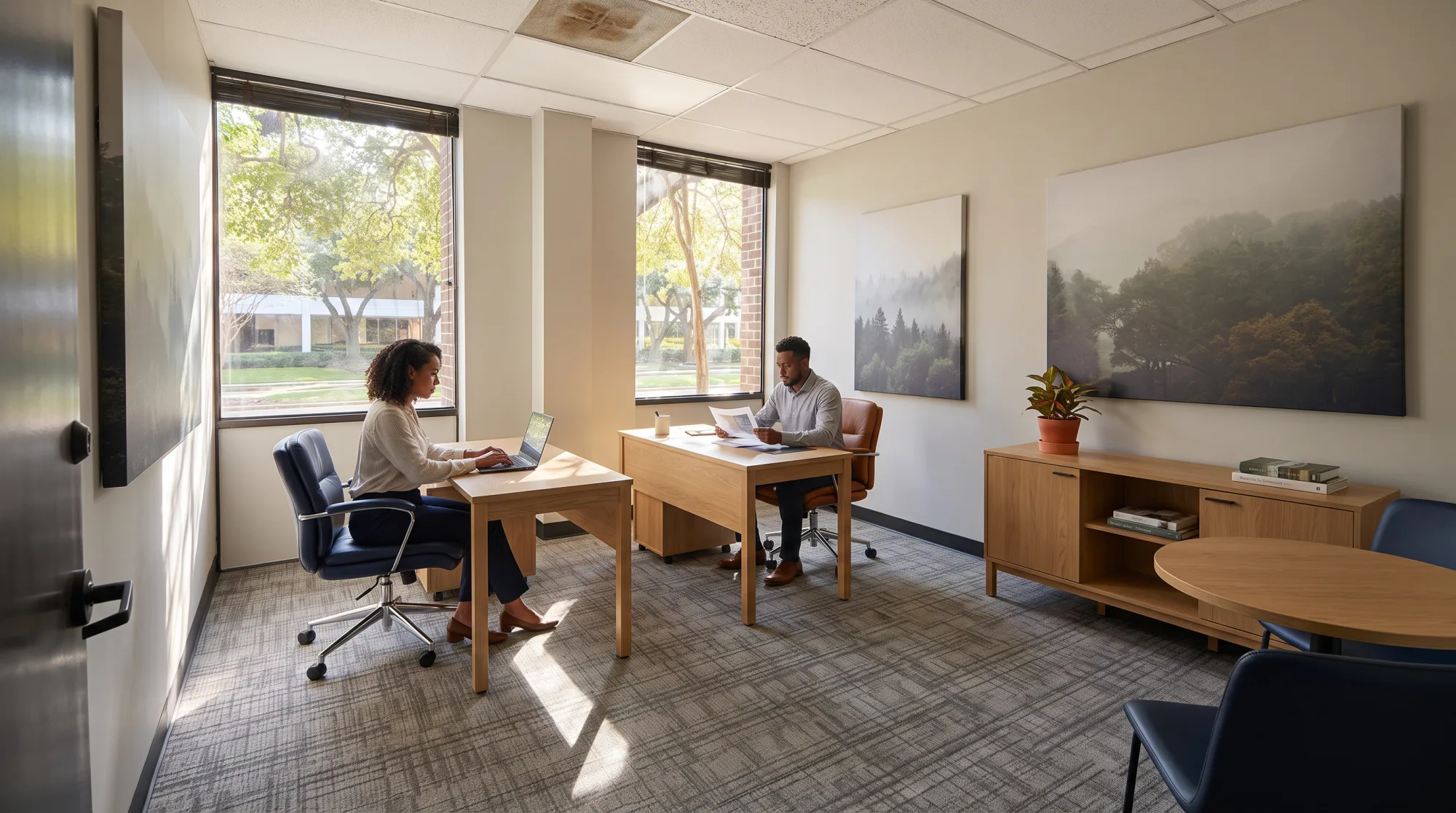 Staged photo of a private office at 1800 St. James Place: two oak desks by the windows, a forest canvas on the wall, two colleagues at work